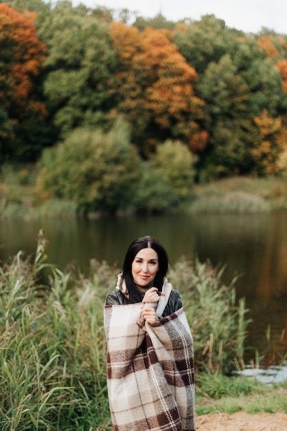 Bella ragazza bruna sorridente si è avvolta in un plaid sullo sfondo della foresta di autunno