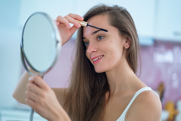 Beautiful smiling brunette woman uses a small round mirror and puts black mascara on her eyelashes during home makeup in the morning