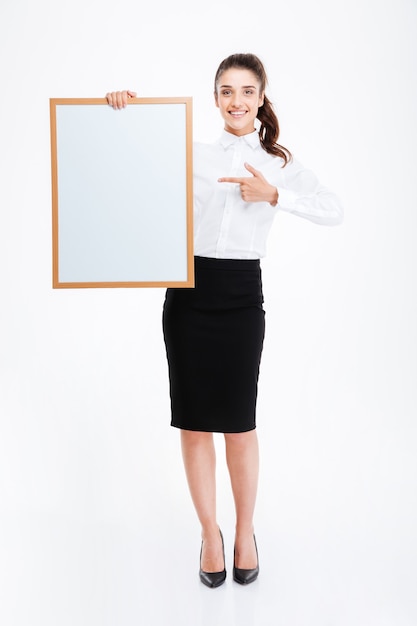 Beautiful smiling businesswoman pointing finger at blank board over white wall