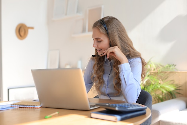 Beautiful smiling call center worker in headphones is working at home office.