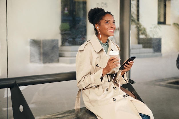 Beautiful smiling casual African American girl in stylish trench coat and earphones with coffee to go happily listening music on cellphone at bus stop