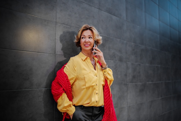 Beautiful smiling caucasian blonde fashionable senior woman with scarf talking on the phone while leaning on the wall outside.