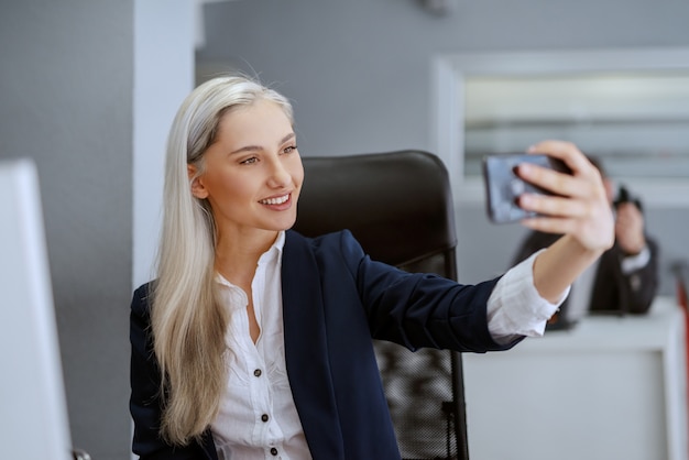 Bella donna bionda caucasica sorridente che si siede nell'ufficio e che la esamina nella riflessione dello smart phone.