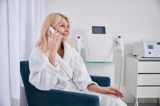 Beautiful smiling Caucasian female in white bathrobe talking on mobile phone in beautician salon