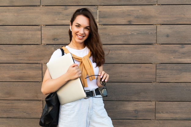 Bella sorridente affascinante giovane donna brunet che guarda l'obbiettivo tenendo il computer portatile e occhiali da sole in t-shirt bianca e jeans azzurri in strada.