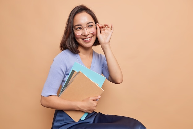 Beautiful smiling clever female student or schoolgirl carries spiral notepads prepares for lectures wears big transparent glasses dressed in neat clothes sits against beige wall