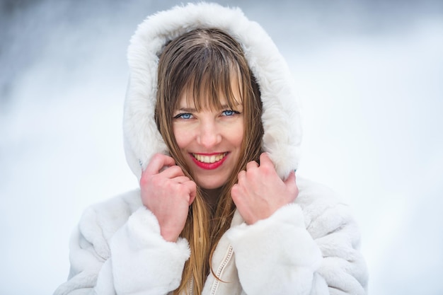 Beautiful smiling confident young white woman pretty face in a\
white fur coat looking at camera posing alone at snowy forest