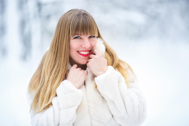 Beautiful smiling confident young white woman pretty face with\
bright red lips in white fur coat looking at camera posing alone at\
snowy forest