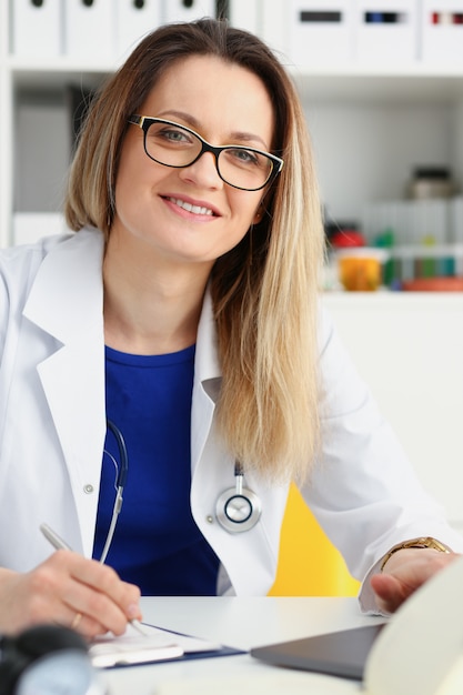 Il bello medico femminile sorridente si siede nel luogo di lavoro. la visita di esame di aiuto al paziente di prevenzione di malattia fisica e 911 intorno al reparto prescrive il concetto di professione del consulente di stile di vita sano del rimedio