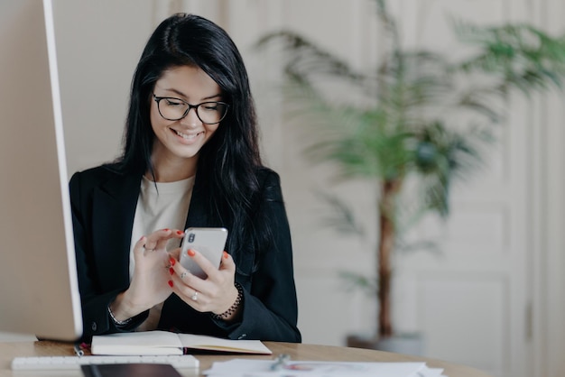 Beautiful smiling female freelancer works remotely concentrated in smartphone sends feedback on received message sits in coworking space dressed formally works on computer surfs internet