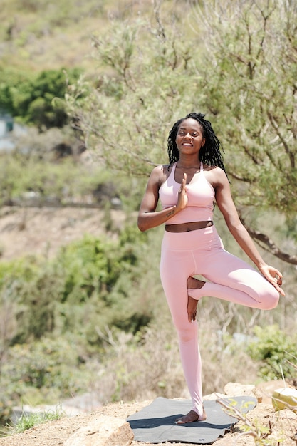 Beautiful smiling fit young woman in legging and sports bra balancing on one leg when doing yoga outdoors