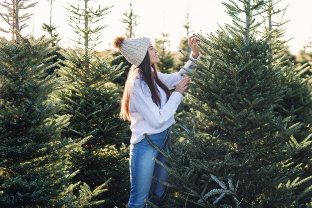 Beautiful smiling girl chooses a Christmas tree on a plantation before winter holidays.