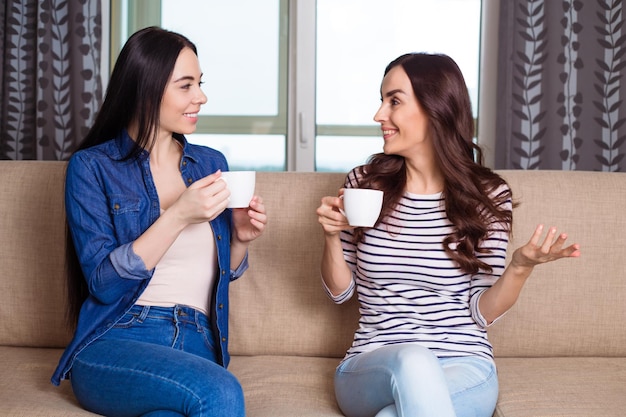 Beautiful smiling girl friends in casual clothes sitting on the sofa at home talking and drinking coffee or tea