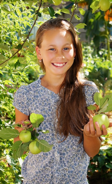 Bella ragazza sorridente nel giardino con mele verdi, vestito blu con fiori