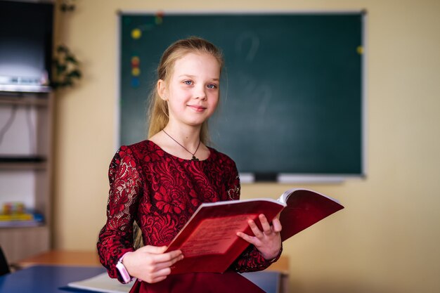 Beautiful smiling girl holding book. Going to school. Portrait, classroom background. Student hugging a book. Lifestyle, interest, hobby, free time, spare time.