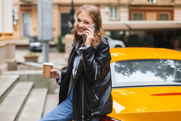 幸せにしながら携帯電話で話している黄色のスポーツカーに寄りかかって行くコーヒーのカップと革のジャケットの美しい笑顔の女の子