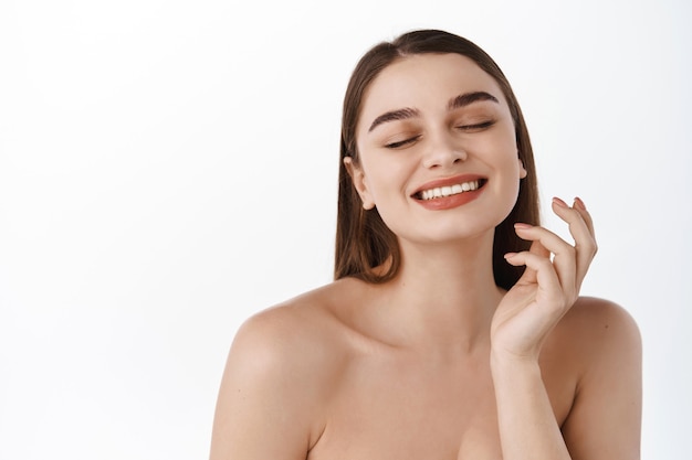 Bellissimo modello di ragazza sorridente con trucco naturale che tocca la pelle idratata incandescente sul muro bianco