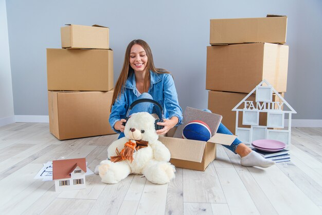 Beautiful smiling girl puts on the headphones on the teddy bear toy and sitting on the floor in the empty new apartment.