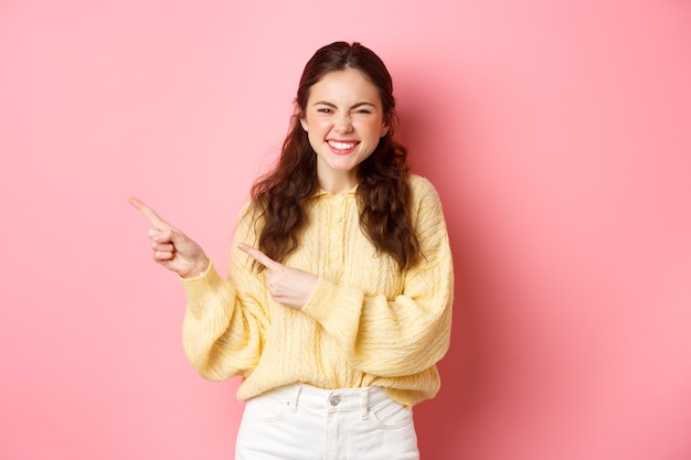 Beautiful smiling girl showing advertisement pointing fingers aside at copy space promoting product on bright pink wall