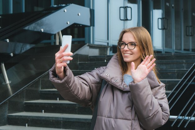 Beautiful smiling girl with glasses in street holding smartphone in hand and greeting with hand