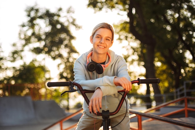 Beautiful smiling girl with headphones happily looking in camera while leaning on bicycle with skatepark on background