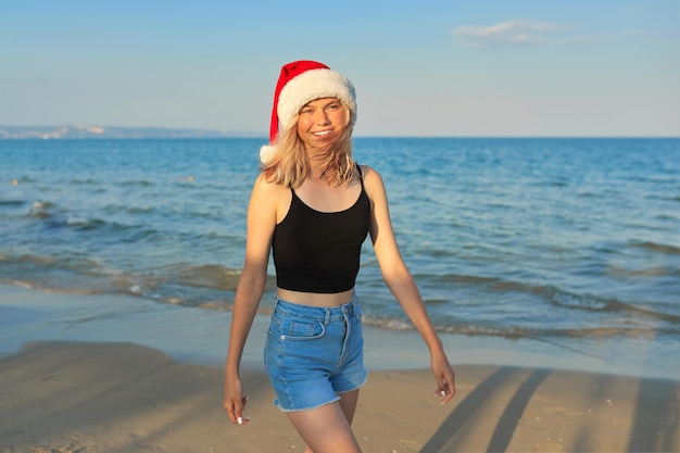 Beautiful smiling happy teenage girl in Santa Claus hat walking along the beach. Christmas and New Year holidays, vacations, travel, tourism concept