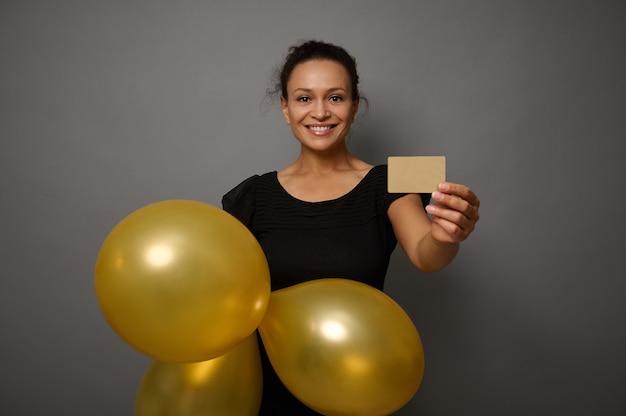 黒の衣装に身を包んだ美しい笑顔の幸せな女性は、金色の膨らんだ気球と金の空の空白のクレジットカードを示しています。灰色の壁の背景にコピー広告スペースとブラックフライデーのコンセプト