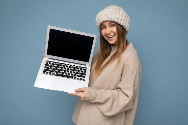 Beautiful smiling happy young dark blond woman in winter warm knitted hat holding computer laptop