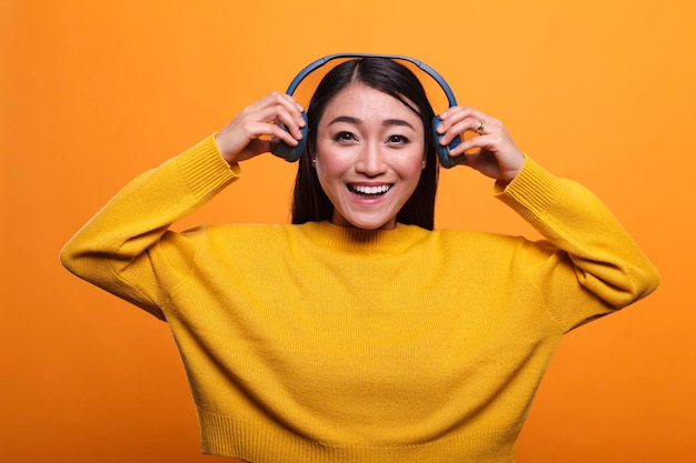 Beautiful smiling heartily woman taking off headphones when she\
meets her friend. cute joyful positive asian person stops listening\
to music to talk with mate while on orange background.