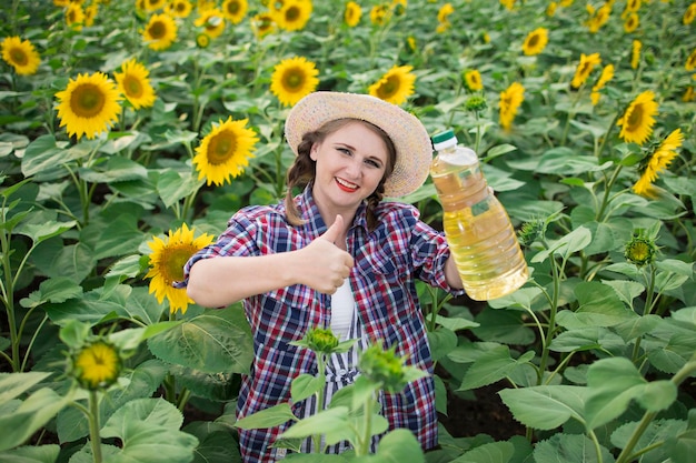 Красивая улыбающаяся радостная женщина-фермер средних лет, держащая в руках бутылки с золотым подсолнечным маслом и поднимающая большой палец вверх на поле урожая подсолнухов в солнечный день