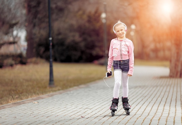 Beautiful smiling little girl inline skating through the park and listening music.