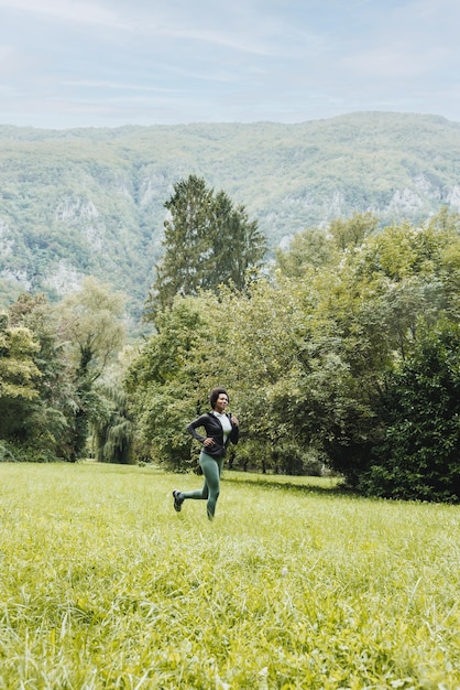 Beautiful smiling mature African American woman is jogging through green grass near the mountain landscape.