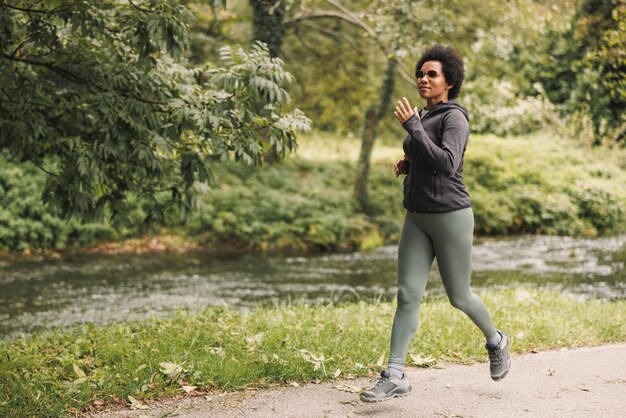 Beautiful smiling mature African American woman is running along trail near the mountains river in nature.