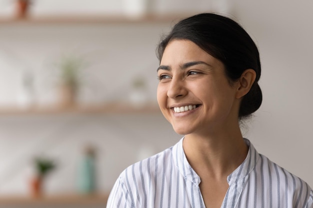 Beautiful smiling millennial indian woman stand indoors look
aside in good optimistic mood think about bright future inspired
excited mixed race female enjoy life dream of positive things copy
space