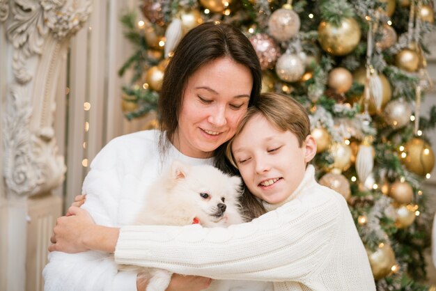 Beautiful smiling mom and son hugging with a happy white dog at home for christmas vacation happy fa...