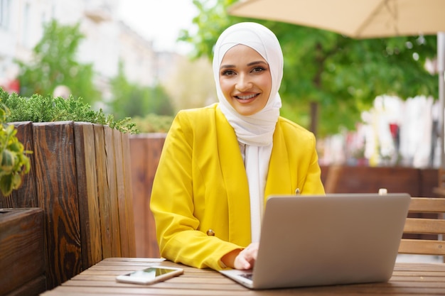 カフェでラップトップを持つ美しい笑顔のイスラム教徒の女性