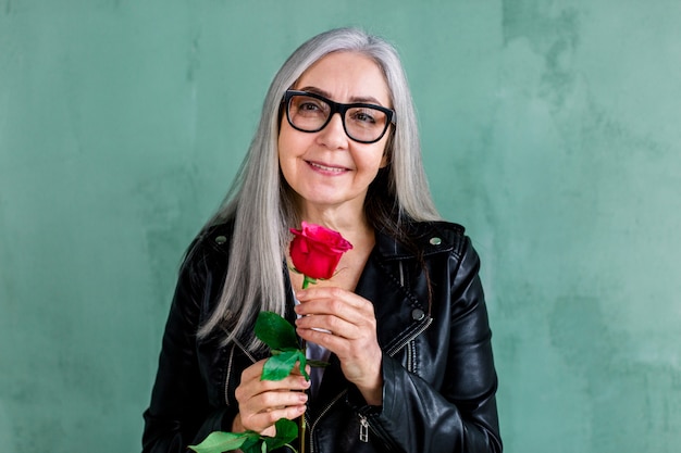 Beautiful smiling senior lady with long straight gray, wearing eyeglasses and stylish leather jacket, posing on camera, standing on green wall background, holding fresh red rose in hand