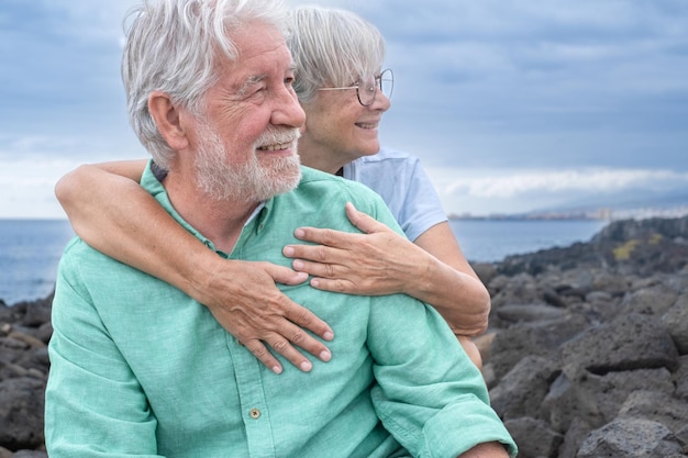 Beautiful smiling whitehaired senior couple sitting together on the cliff looking away Cloudy sky in background copy space