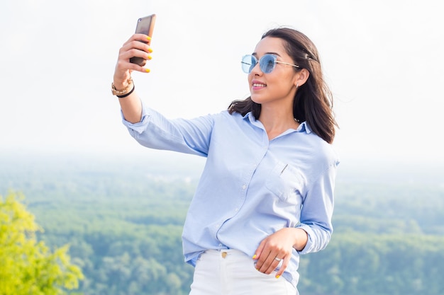 Bella donna sorridente con gli occhiali blu si fa selfie nella natura in montagna