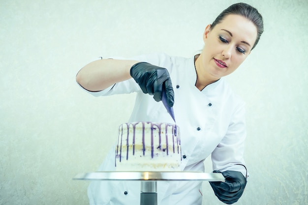 Красивая и улыбающаяся женщина-кондитер в черных перчатках и белой рабочей форме украшает торт на кухне. кондитер, торт, кулинария. свадебный и праздничный торт с ягодами.