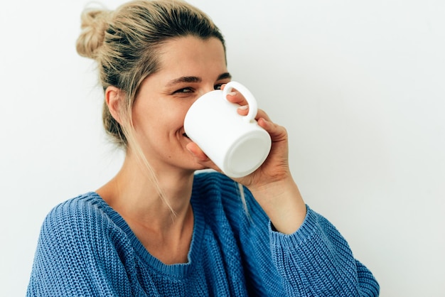 Beautiful smiling woman drinking cappuccino Happy young woman drinking hot beverage at home Charming blonde female holding a coffee cup Lady wearing blue sweater sitting with cup of tea in her hand