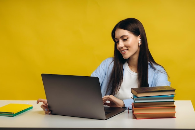 Beautiful smiling woman sitting at table using laptop and writing in her notebook on yellow background Education concept