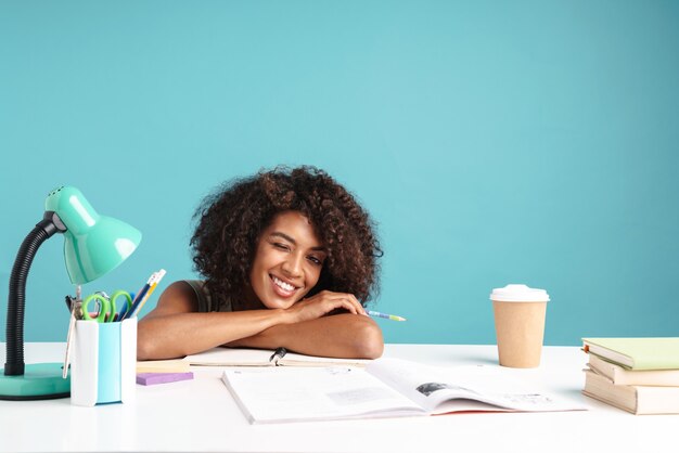 Beautiful smiling young african businesswoman casually dressed laying on a desk isolated over blue wall