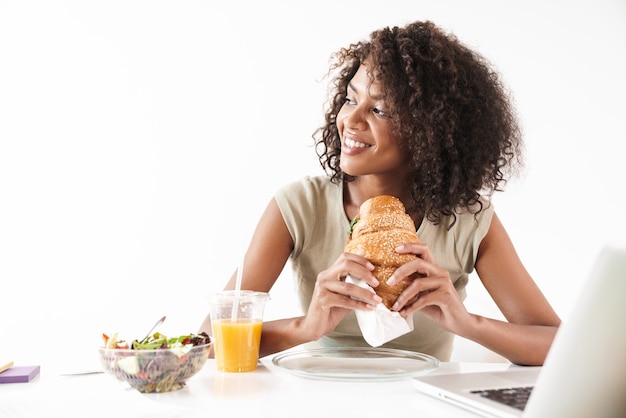 写真 白い壁に隔離された机に座って、ラップトップコンピューターで作業しながら昼食をとって、美しい笑顔の若いアフリカの女性