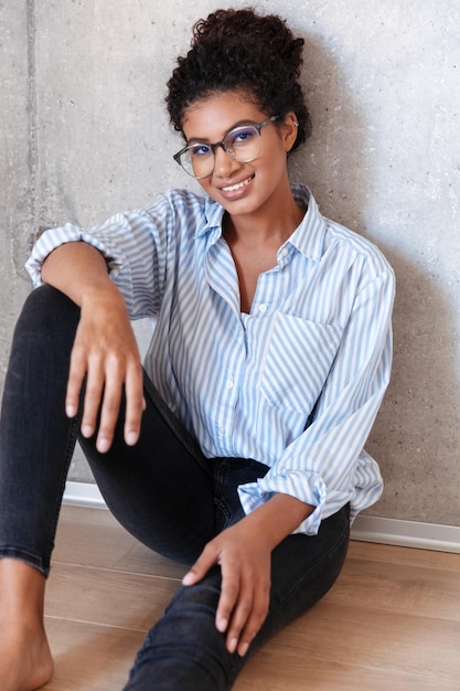 Beautiful smiling young african woman wearing casual clothes and glasses leaning on a wall indoors, posing while sitting