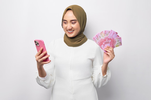 Beautiful smiling young Arabian Asian Muslim woman wearing hijab holding cash money in rupiah banknote and mobile phone isolated on white background People religious lifestyle concept