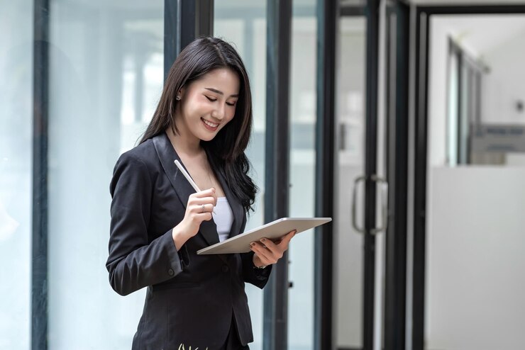 Photo beautiful smiling young asian businesswoman standing holding a pen using a  tablet happy successful at the office.