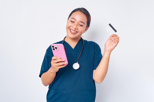 青い制服を着て携帯電話を使用し、白い背景に分離されたクレジット バンク カードを保持している美しい笑顔の若いアジア女性専門の看護師