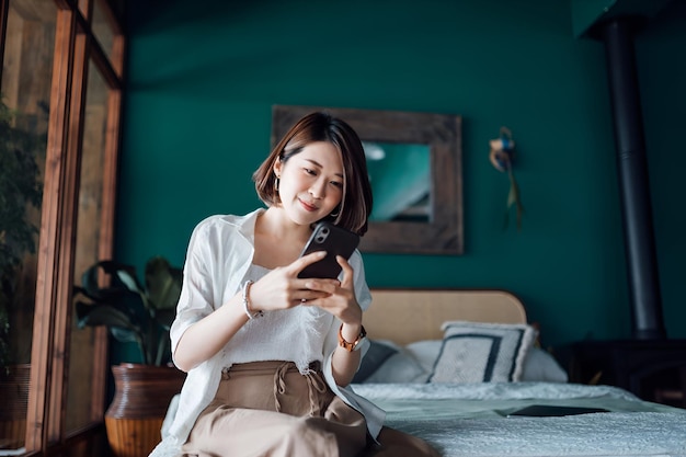 Beautiful smiling young Asian woman sitting on her bed text messaging on smartphone at home Technology in everyday life