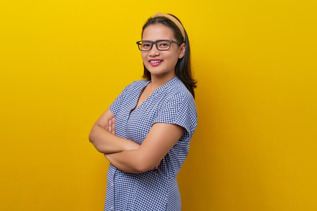 Beautiful smiling young asian woman wearing a dress checkered with glasses crossed hands and folded looking confident on camera isolated on yellow background people lifestyle concept